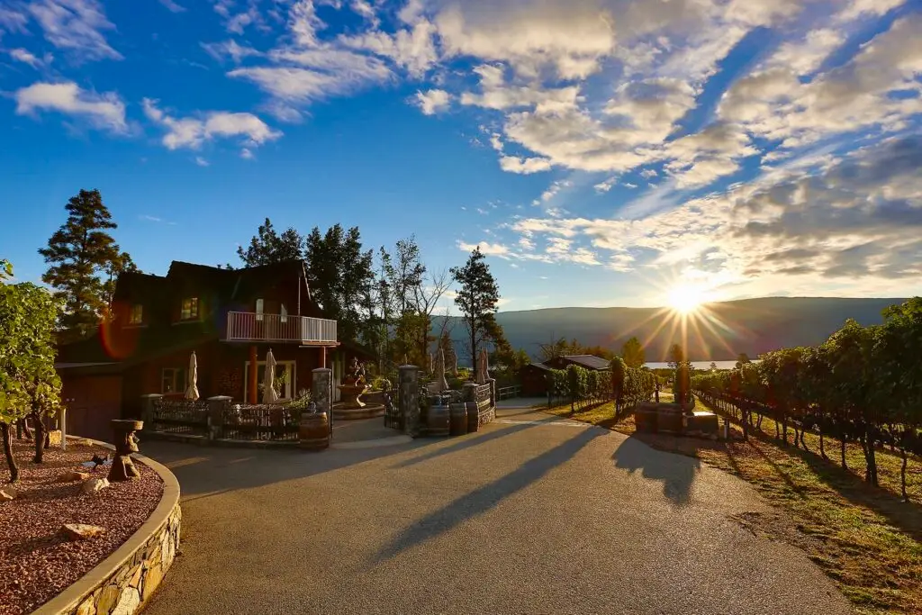 A view of Heaven's Gate winery. The tasting room is pictured in a beautiful 2 story log home, nestled int he vineyards overlooking Okanagan Lake. The morning sun is peaking over the eastern mountains on the other side of the lake, with bright blue sky. The setting is in early fall, with leaves still on the vines, but with leaves starting to fall on the tall beautiful trees surrounding the winery.